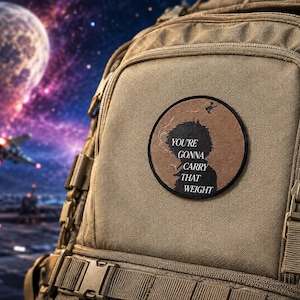 May include: A circular embroidered patch featuring a black silhouette of a person with a cigarette and smoke, set against a brown background. The text reads, "YOU'RE GONNA CARRY THAT WEIGHT." The patch is attached to a tan tactical backpack.