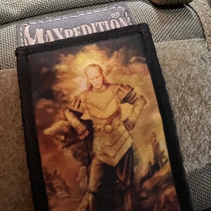 May include: A black and white patch with the text "Maxpedition" in white. The patch features a painting of a person in armour standing in a field with a sword in their hand.