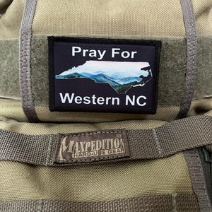 Op de afbeelding: Een bruine Maxpedition Hard Use Gear rugzak met een zwarte patch waarop "Pray For Western NC" staat en een blauwe en witte omtrek van de staat North Carolina.