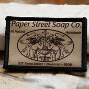 May include: Black and white woven patch with the text "Paper Street Soap Co." in white lettering. The patch also includes the text "All Natural" and "Handmade" in smaller white lettering. The patch features a design of two angels holding a bowl with a fountain in the centre.