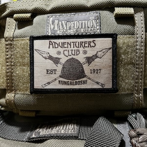 May include: A brown and black patch with the text "Adventurers Club Est 1927 Kungaloosh!" with a graphic of two spears and a helmet.