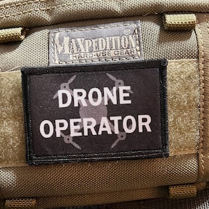May include: A black patch with white text that reads "DRONE OPERATOR" and a stylized drone graphic. The patch is attached to a brown tactical bag with a black Maxpedition Hard-Use Gear label.