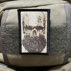 May include: A black and white embroidered patch featuring a skull with a long beard. The patch is sewn onto a brown fabric background.
