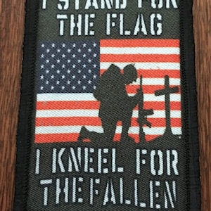 May include: A black and green patch with the text "I stand for the flag I kneel for the fallen" and a silhouette of a soldier kneeling in front of a cross with the American flag in the background.