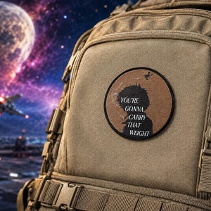 May include: A circular embroidered patch attached to a tan tactical backpack. The patch features a brown background with a black silhouette of a person smoking, accompanied by the text "YOU'RE GONNA CARRY THAT WEIGHT" in a serif font.