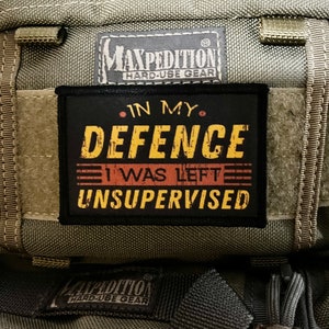 May include: A black patch with yellow text that reads "In My Defence I Was Left Unsupervised" attached to a green Maxpedition bag with the brand name and "Hard-Use Gear" printed on it.