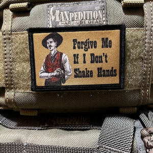 Puede incluir: Un parche de color beige y negro con un vaquero de dibujos animados y el texto "Forgive Me If I Don't Shake Hands" sobre un fondo beige. El parche está unido a una bolsa de color beige y negro.