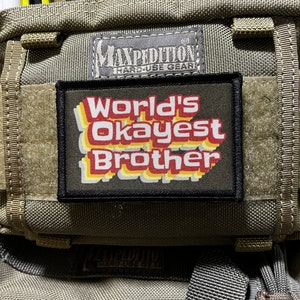 Puede incluir: Un parche negro y verde con el texto "World's Okayest Brother" en una fuente retro con colores rojo, amarillo y naranja. El parche está unido a una bolsa táctica verde con el logotipo de Maxpedition.