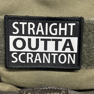 Puede incluir: Parche blanco y negro con el texto "STRAIGHT OUTTA SCRANTON" en letras blancas sobre fondo negro.