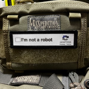 Puede incluir: Una bolsa verde Maxpedition con un parche negro que dice "No soy un robot" y un logotipo de reCAPTCHA. La bolsa tiene una cremallera y una correa.