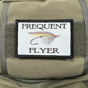 May include: A black and white patch with the text "FREQUENT FLYER" in black lettering. A stylised image of a fly fishing lure is centred above the text.
