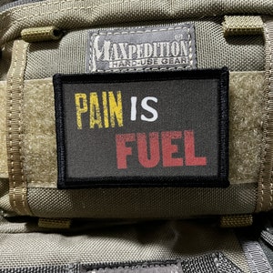 May include: A black patch with white text that reads "Pain is Fuel" in yellow and red letters. The patch is attached to a beige Maxpedition bag with the brand name and "Hard-Use Gear" printed on it.
