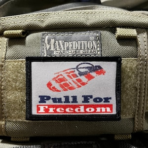 Può includere: Una borsa verde Maxpedition Hard-Use Gear con una toppa nera che dice "Pull For Freedom" e una grafica rosso, bianco e blu di una linguetta.