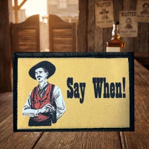 May include: A rectangular embroidered patch with a black border on a light yellow background. It features a graphic of a person wearing a black hat and a red vest, with the black text "Say When!" printed to the right of the figure.
