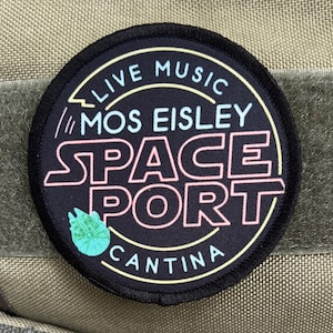 May include: A black round patch with a white outline. The text "Live Music // Mos Eisley Space Port Cantina" is written in a retro font with pink, blue, and yellow accents. A green alien creature is in the lower left corner.