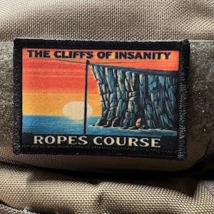 Könnte beinhalten: Ein schwarz-weißes Patch mit einer Sonnenuntergangsszene und einer Felswand. Der Text "THE CLIFFS OF INSANITY" befindet sich oben und "ROPES COURSE" unten.