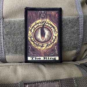 Puede incluir: Un parche bordado negro y marrón con un ojo amarillo y el texto "The Ring" sobre un fondo beige.