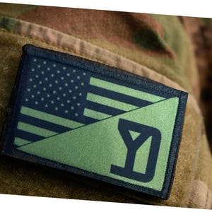 Op de afbeelding: Een rechthoekige patch met een gesplitst ontwerp: de bovenste helft toont de Amerikaanse vlag, terwijl de onderste helft een groene achtergrond heeft met de donkerblauwe letter "Y". De patch is bevestigd op een camouflage stof.