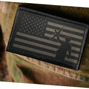 May include: A rectangular patch featuring the American flag in shades of black and olive green. A silhouette of a person holding a rifle aloft is superimposed on the flag. The patch is affixed to a camouflage fabric.
