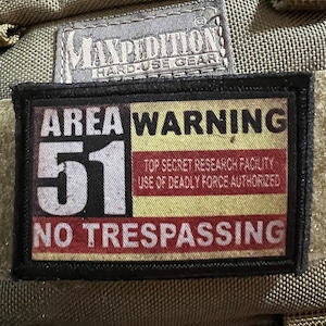 Puede incluir: Un parche rectangular con el texto "AREA WARNING 51 NO TRESPASSING" en letras blancas y rojas. El parche también incluye el texto "TOP SECRET RESEARCH FACILITY USE OF DEADLY FORCE AUTHORIZED". El parche es negro con fondo beige.