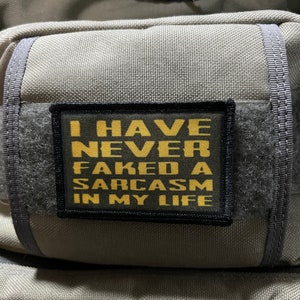 May include: A black and yellow patch with the text "I HAVE NEVER FAKED A SARCASM IN MY LIFE" on a gray fabric background.