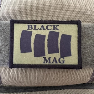 May include: A rectangular patch with the words "BLACK MAG" in white block letters. Below the word "BLACK" are four dark blue vertical rectangles. The patch is on a green background with a black border. The background is a tan fabric.