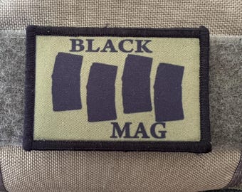 Può includere: Una toppa rettangolare con le parole "BLACK MAG" in lettere bianche. Sotto la parola "BLACK" ci sono quattro rettangoli verticali blu scuro. La toppa è su uno sfondo verde con un bordo nero. Lo sfondo è un tessuto color cachi.