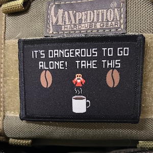 May include: Black and white embroidered patch with the text "It's dangerous to go alone! Take this" and a pixelated image of a coffee cup with steam rising from it, two coffee beans on either side of the cup, and a pixelated image of a person holding a coffee cup.