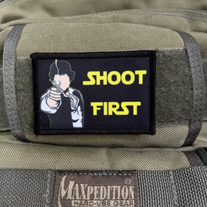 Puede incluir: Un parche negro con el texto amarillo "SHOOT FIRST", que representa una figura de dibujos animados sosteniendo un arma. El parche está adherido a una mochila beige.
