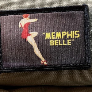 May include: A black patch with a colorful image of a woman in a red dress and the text "Memphis Belle" in yellow.