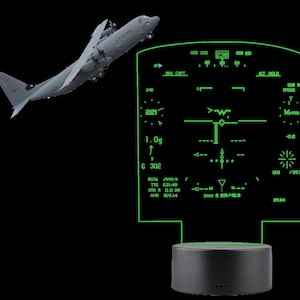 Può includere: Un aereo militare grigio in volo, con un pannello strumenti di volo verde illuminato in primo piano. Il pannello mostra vari dati di volo, tra cui altitudine, velocità e rotta. La base è un cilindro grigio scuro.
