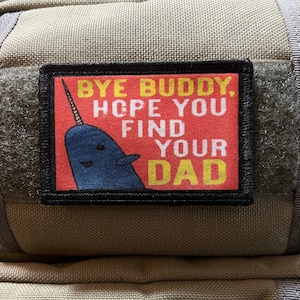 May include: A rectangular patch with the text "BYE BUDDY. HOPE YOU FIND YOUR DAD" in yellow and white on a red background. A blue narwhal illustration is featured. The patch is bordered in black and attached to a tan fabric.