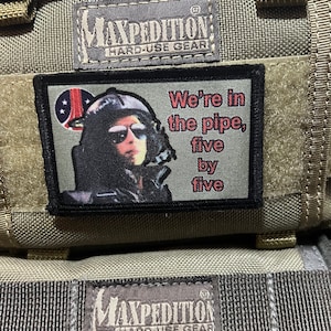 May include: A green tactical bag with two black patches. The top patch says "MAXPEDITION HARD-USE GEAR". The bottom patch features a cartoon image of a person wearing a helmet and sunglasses with the text "We're in the pipe, five by five".