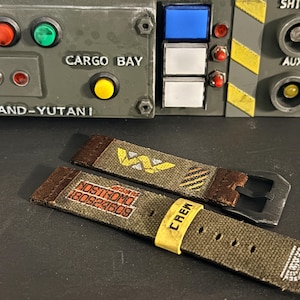 May include: Two olive green canvas watch straps with brown leather ends and a black buckle. One strap features a yellow Weyland-Yutani logo and hazard stripes. The other strap displays the text NOSTROMO 180824609 and a yellow CREW loop.