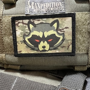 May include: A green tactical bag with a black patch featuring a cartoon raccoon with red eyes. The patch is labelled "Maxpedition Hard-Use Gear".