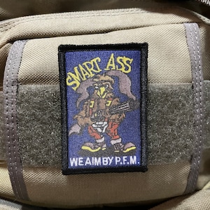 May include: A black and blue patch with a cartoon bird wearing a red and white outfit and holding a gun. The patch says "SMART ASS" and "WE AIM BY P.F.M."