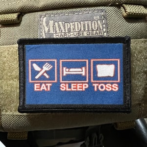 May include: A blue patch with white and red text that reads "Eat Sleep Toss". The patch has three white squares with icons inside. The first square has a fork and knife, the second square has a bed, and the third square has a pillow. The patch is attached to a green bag with a black label that reads "Maxpedition Hard-Use Gear".