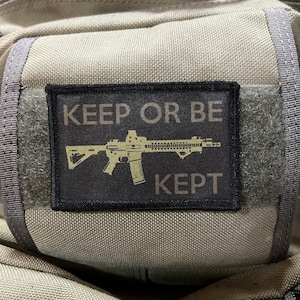 May include: A black patch with white text that reads "KEEP OR BE KEPT" and a silhouette of a rifle in the center. The patch is attached to a gray fabric bag.