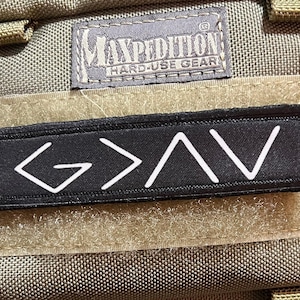 Puede incluir: Un parche rectangular negro con el texto blanco G > ^ V, sujeto a una superficie de tela color canela Maxpedition hard-use gear con un cierre de velcro. El parche está colocado horizontalmente debajo de una etiqueta de marca gris Maxpedition.