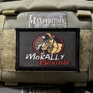 May include: A black patch with white text that reads "Morally Flexible" and a red circle with a cartoon image of a man in a fedora and trench coat. The patch is attached to a green tactical bag with a black label that reads "Maxpedition Hard-Use Gear".