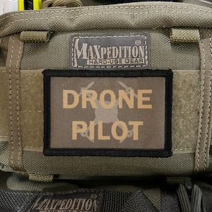 May include: A green Maxpedition bag with a black patch that says "Drone Pilot" in yellow letters. The bag has a black strap and a Maxpedition logo on the front.