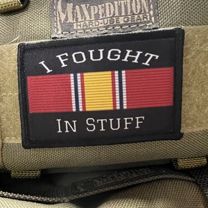 Puede incluir: Un parche negro con texto blanco que dice "I FOUGHT IN STUFF" y una cinta roja, blanca y amarilla. El parche está unido a una bolsa táctica verde con una etiqueta negra que dice "MAXPEDITION HARD-USE GEAR".