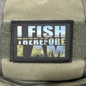 Puede incluir: Un parche rectangular con el texto "I FISH THEREFORE I AM" en letras blancas. El fondo del texto es un paisaje con cielo azul, agua y hierba verde. El parche está sobre una tela verde.