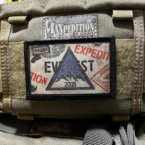 May include: A black and white patch with a blue triangle and the text "EXPEDITION" in red. The triangle has a mountain range inside and the text "2021" below it. The patch is attached to a brown and green bag.