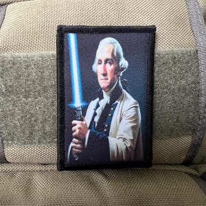 May include: A black and white patch with a portrait of a man holding a blue lightsaber. The man is wearing a white shirt and a black jacket. The patch is on a brown background.