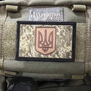 May include: A green military-style backpack with a black patch featuring the Ukrainian coat of arms, a trident, on a camouflage background. The patch is bordered in black and has the text "Maxpedition Hard-Use Gear" above it.