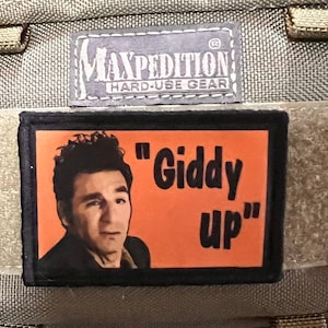 Könnte beinhalten: Ein schwarzer und orangefarbener Patch mit einem Bild eines Mannes mit braunen Haaren und einem schwarzen Hemd. Der Text auf dem Patch lautet "Giddy Up".