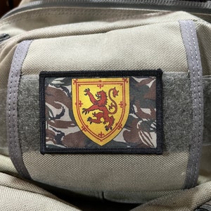 Op de afbeelding: Een beige rugzak met een zwarte patch met daarop de Schotse leeuw op een geel schild met rode accenten. De patch is op een camouflage achtergrond.