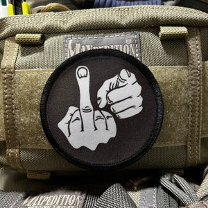 Puede incluir: Un parche blanco y negro con un dedo medio y un puño en una mochila verde y marrón.