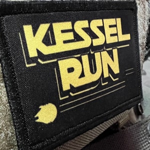 Peut inclure: Un patch noir avec du texte jaune qui dit "Kessel Run" avec une image stylisée jaune du Faucon Millenium dans le coin inférieur gauche.